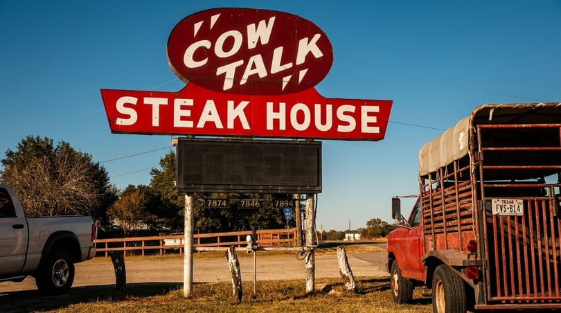 Red "Cow Talk Steak House" sign with a blank marquee, a silver pickup, and a red truck with a cattle trailer.