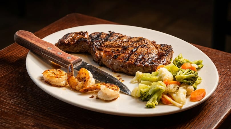 Grilled steak, shrimp, and mixed vegetables with a steak knife on a white plate.