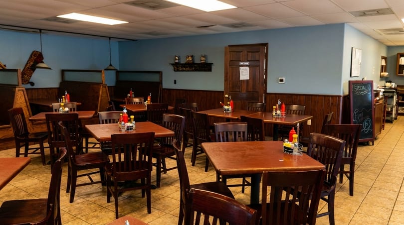 An empty diner interior with wooden tables, chairs, and booths, light blue walls, and tiled floors.